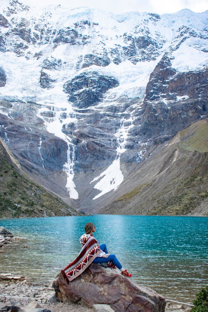 A serene view of a person in poncho sitting by Humantay Lake in Cusco, Peru, surrounded by snowy mountains.